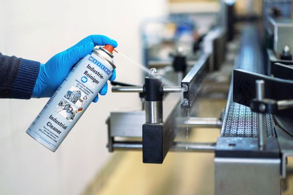 Ruka v modré rukavici aplikuje sprej WEICON Industrie-Reiniger na kovový díl průmyslového dopravníku.