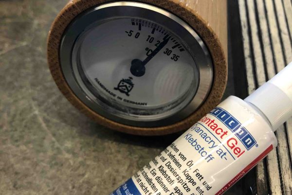 Aanalogový teploměr v dřevěném pouzdře ukazující přibližně 23 °C vedle tuby lepidla WEICON Contact Gel.