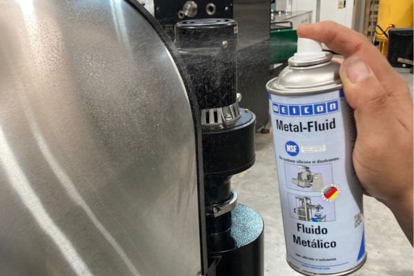 Ruka stříká sprej WEICON Metal-Fluid na zakřivený nerezový povrch průmyslového stroje.