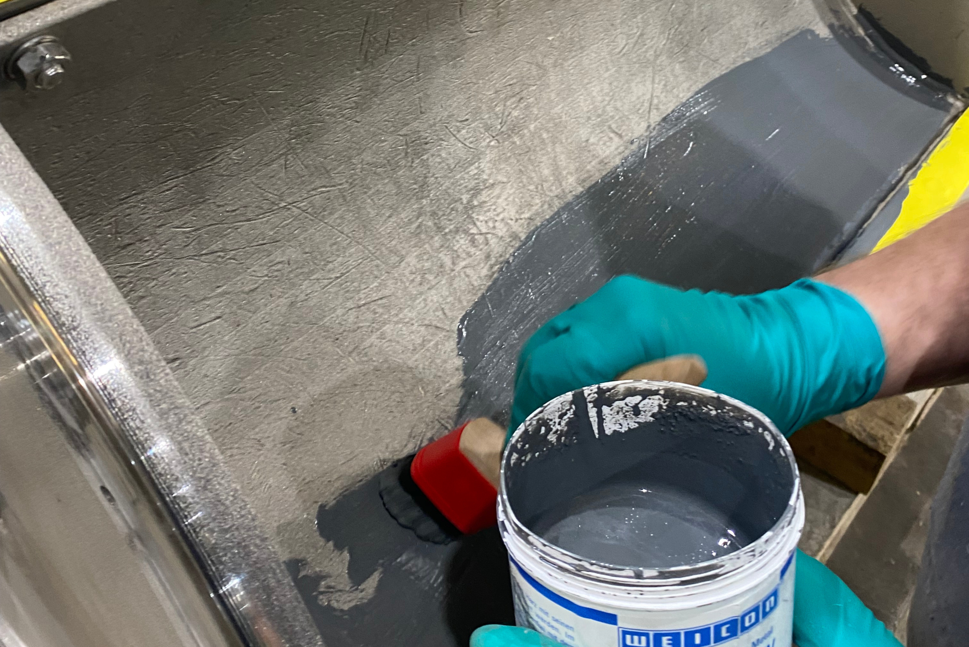 Detailní pohled na pracovníka v zelených ochranných rukavicích, který pomocí štětce nanáší šedou ochrannou vrstvu ze štítkem označené nádoby na vnitřní kovový povrch zařízení.