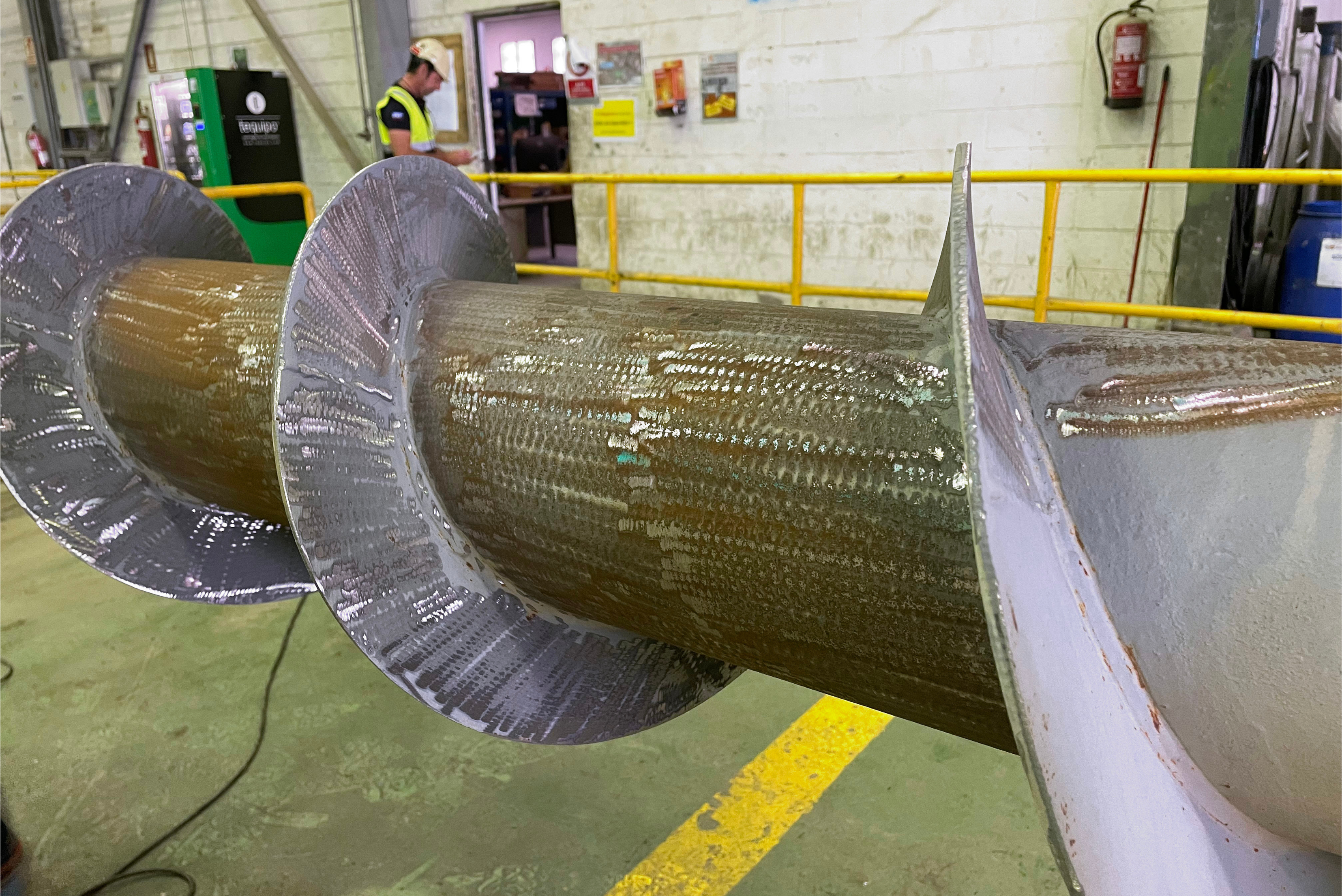 Detailní pohled na průmyslovou šnekovou hřídel (dopravní šnek) s ošetřeným a natřeným povrchem v industriálním prostředí.