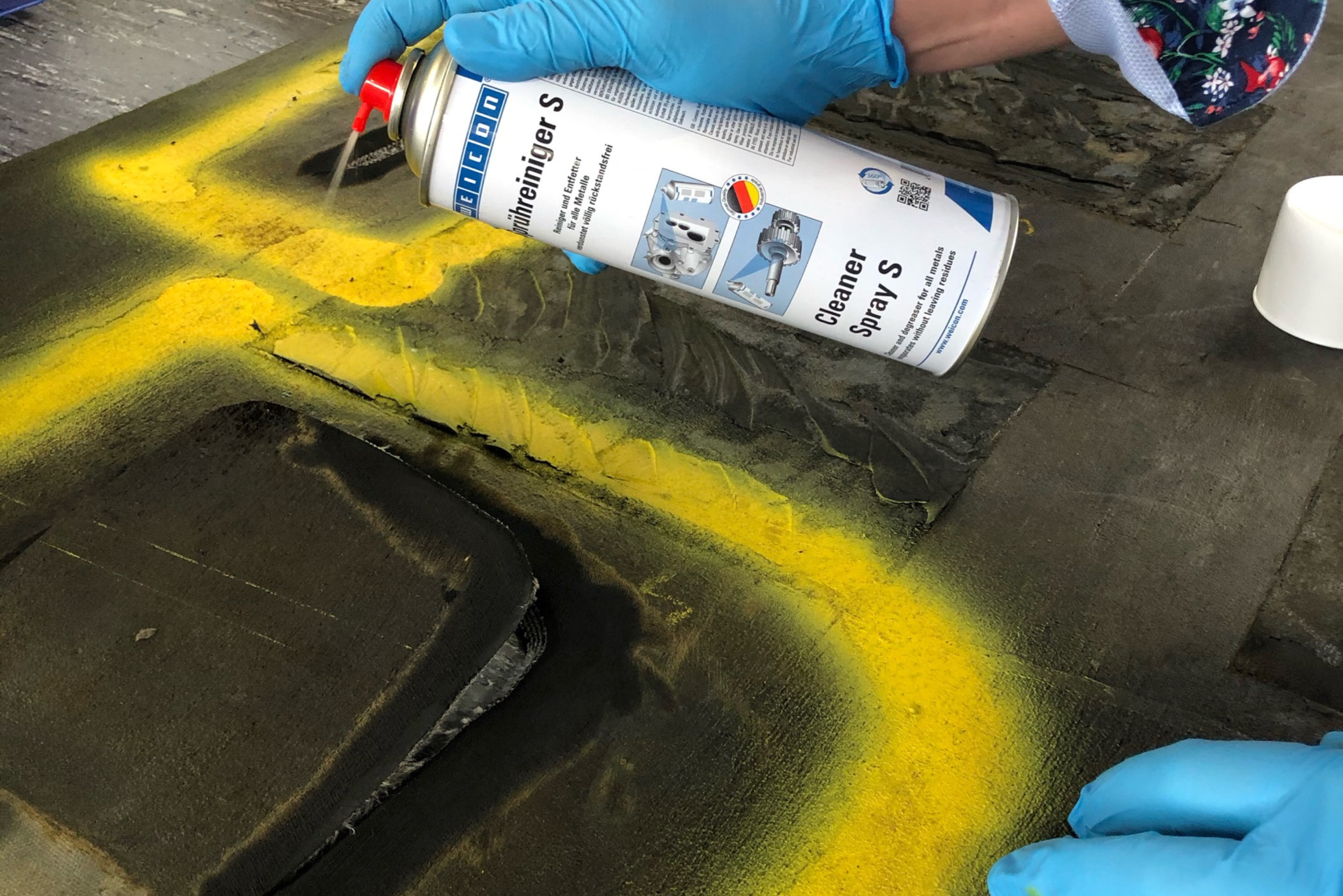 Close-up view of a person wearing blue protective gloves spraying a canister of WEICON Cleaner Spray S onto a dark material surface marked with yellow lines.